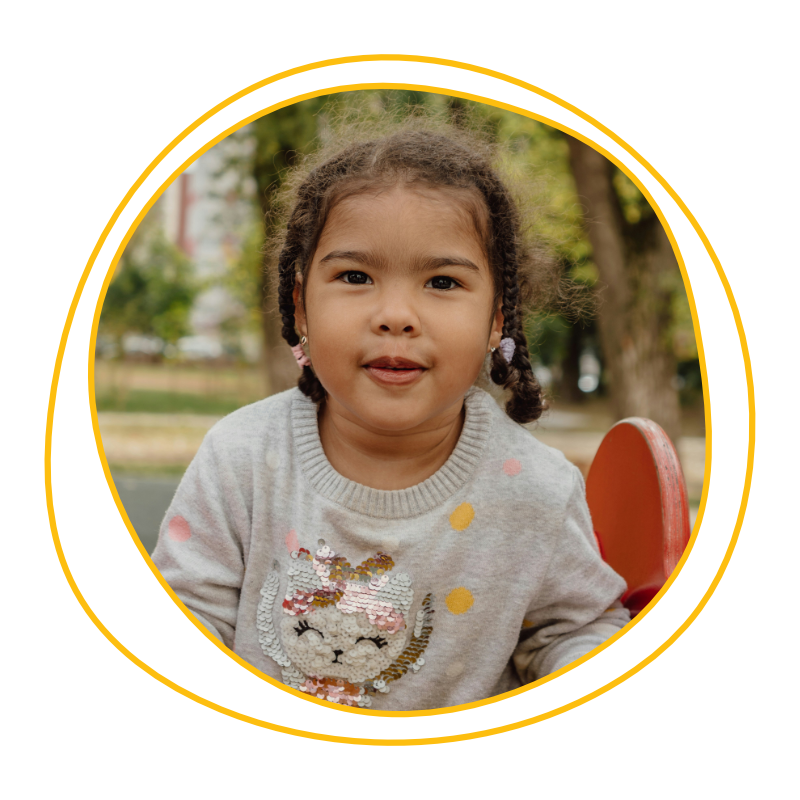 Young girl with braids, wearing a sweater with a sequined cat design, sits outdoors. She's framed by trees, creating a playful and warm atmosphere.