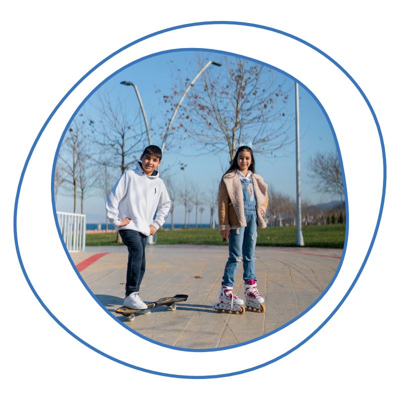 Two kids outdoors, one on a skateboard, the other on rollerblades. They smile under a clear blue sky, surrounded by leafless trees and streetlights.