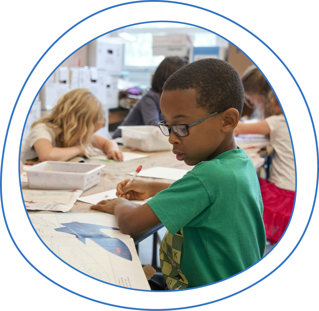 A young boy in glasses focuses on his drawing at a classroom table, surrounded by other children engaged in their creative work